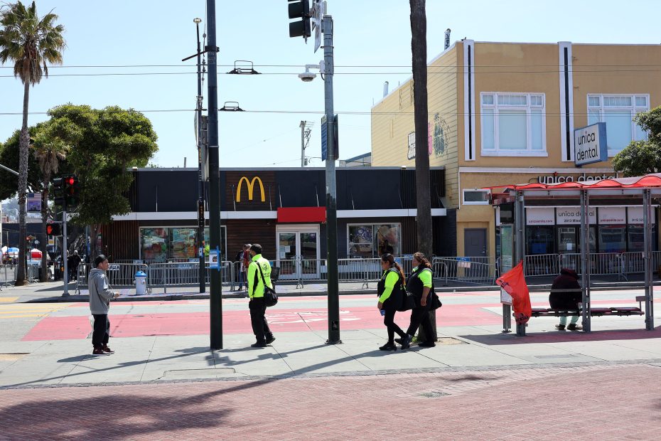 Several people, including workers in yellow vests, walk and wait at a street intersection in front of a McDonald's and other businesses on a sunny day.
