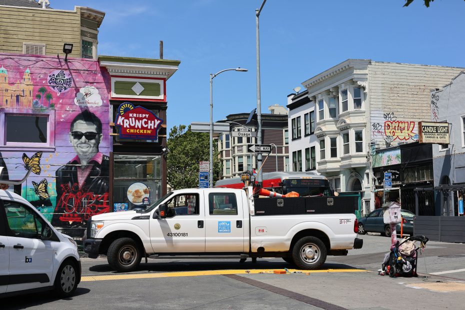 A white utility truck drives through an urban intersection with street art, shops, and a Krispy Krunchy Chicken sign in the background. A stroller sits unattended on the sidewalk.