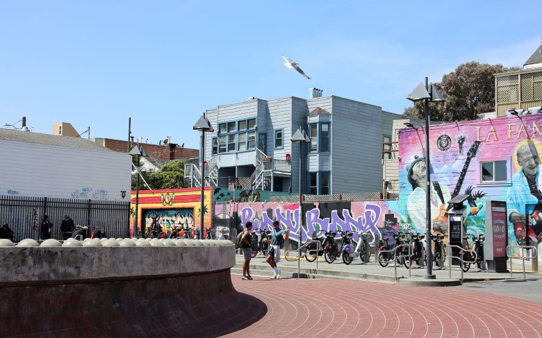 Outdoor urban scene with colorful graffiti murals, a row of parked bikes, people near a fence, and a seagull flying above blue and gray buildings under a clear sky.