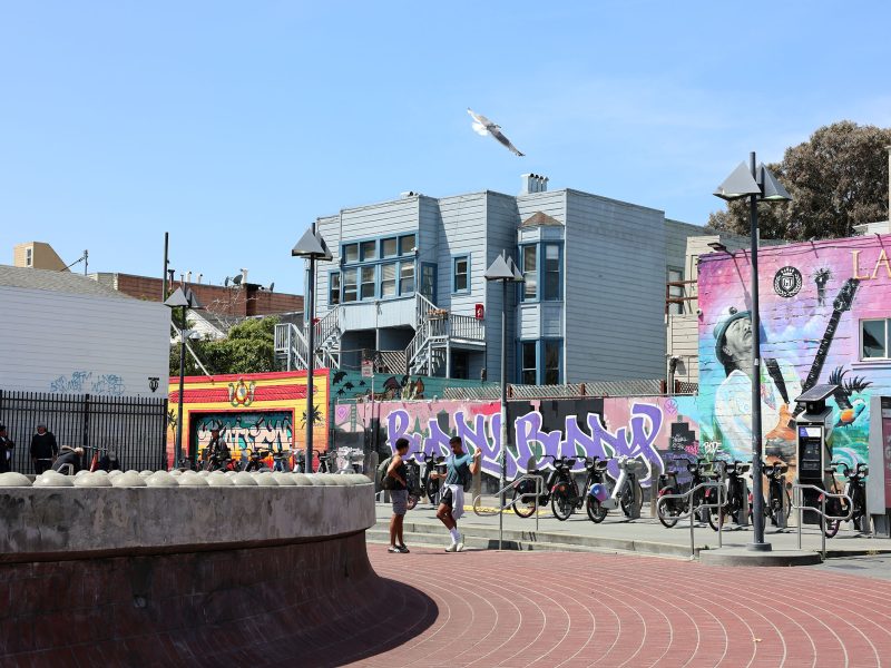 Outdoor urban scene with colorful graffiti murals, a row of parked bikes, people near a fence, and a seagull flying above blue and gray buildings under a clear sky.