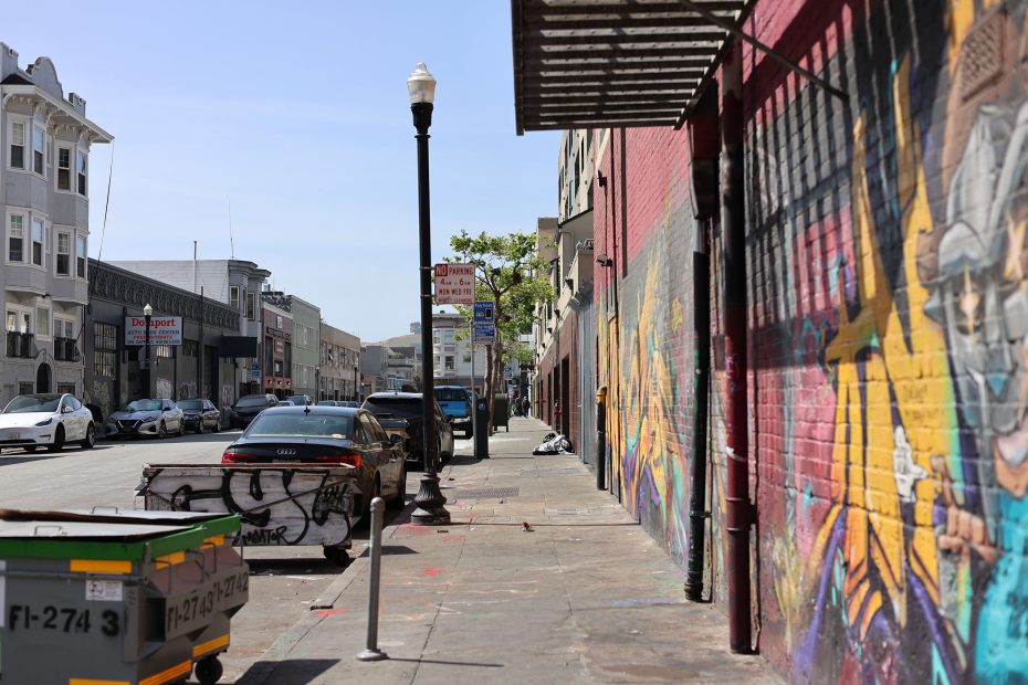 A city street with parked cars, a colorful mural on a brick wall, a dumpster, and a lamp post on the sidewalk under a clear sky.