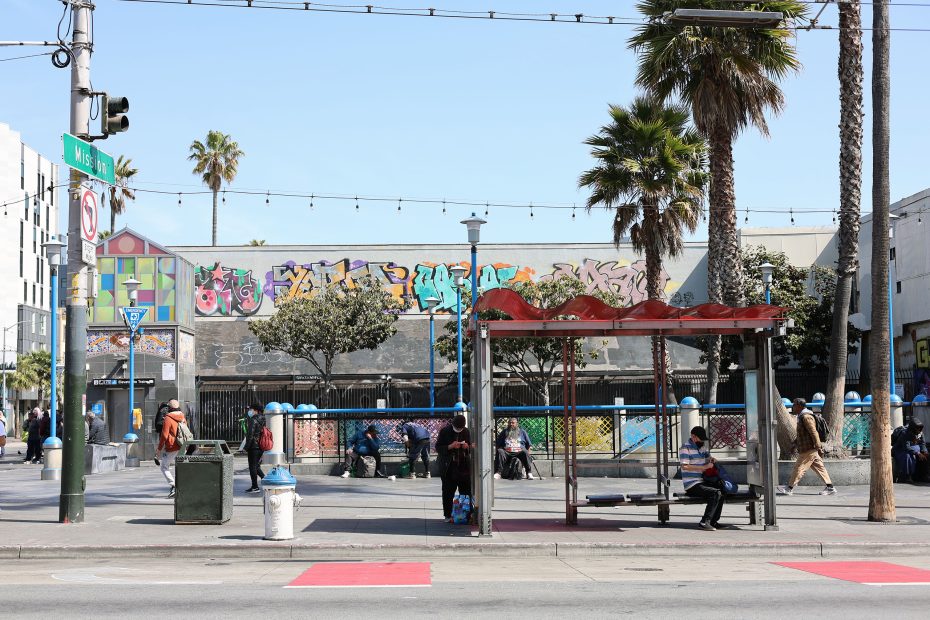 A city bus stop with people sitting and standing, palm trees, street art on a building, and urban street elements visible.