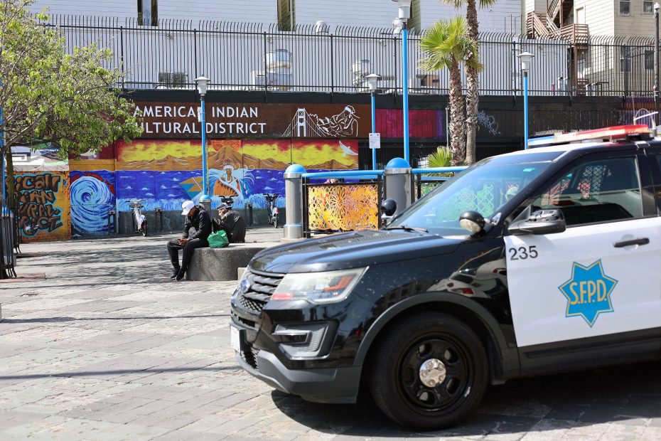 A San Francisco police car is parked near a mural reading "American Indian Cultural District" while people sit on benches in the background.