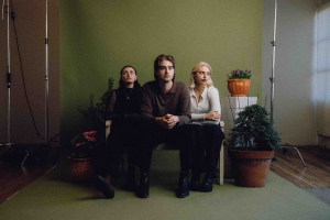 Three people sit on a bench indoors, surrounded by potted plants, against a green background.