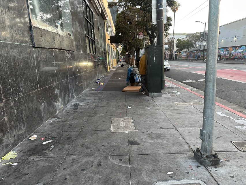 A city sidewalk with scattered litter, a person standing beside belongings near a building, and graffiti visible in the background.