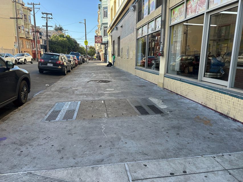 A city sidewalk with parked cars on the left and buildings on the right; one person is sitting alone near the end of the block.