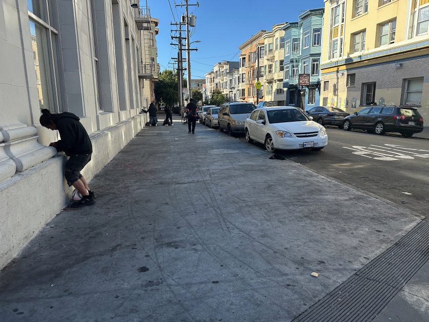 A city sidewalk with people standing and walking, cars parked along the street, and residential buildings in the background on a clear day.