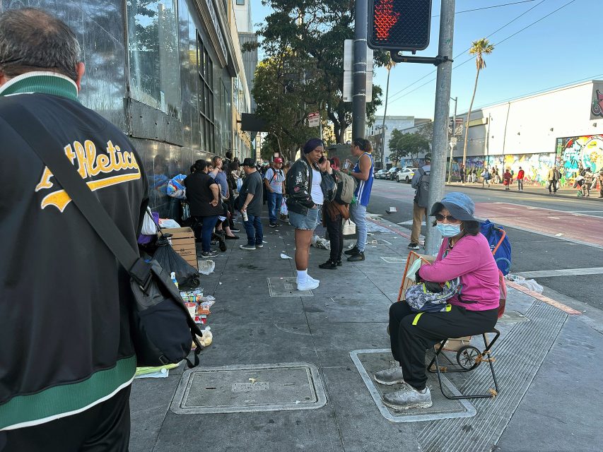 People line up on a city sidewalk next to a street, with some sitting and others standing; various items are placed on the ground nearby.