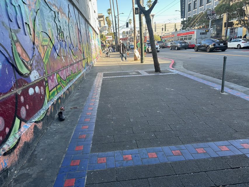 A city sidewalk with a colorful mural on the left wall, blue and red tiles bordering the pavement, and people walking in the background near a street with cars and a tram.