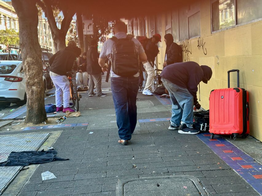 Several people stand and bend down on a city sidewalk next to a yellow wall, with scattered belongings and a red suitcase nearby.
