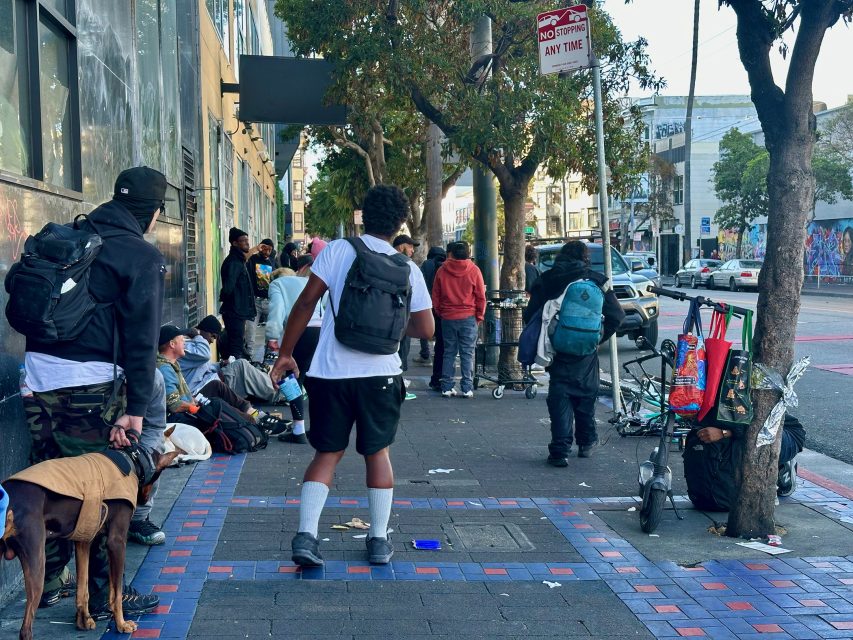A group of people, some with backpacks and belongings, gather on a city sidewalk near a building; a dog stands with one person.