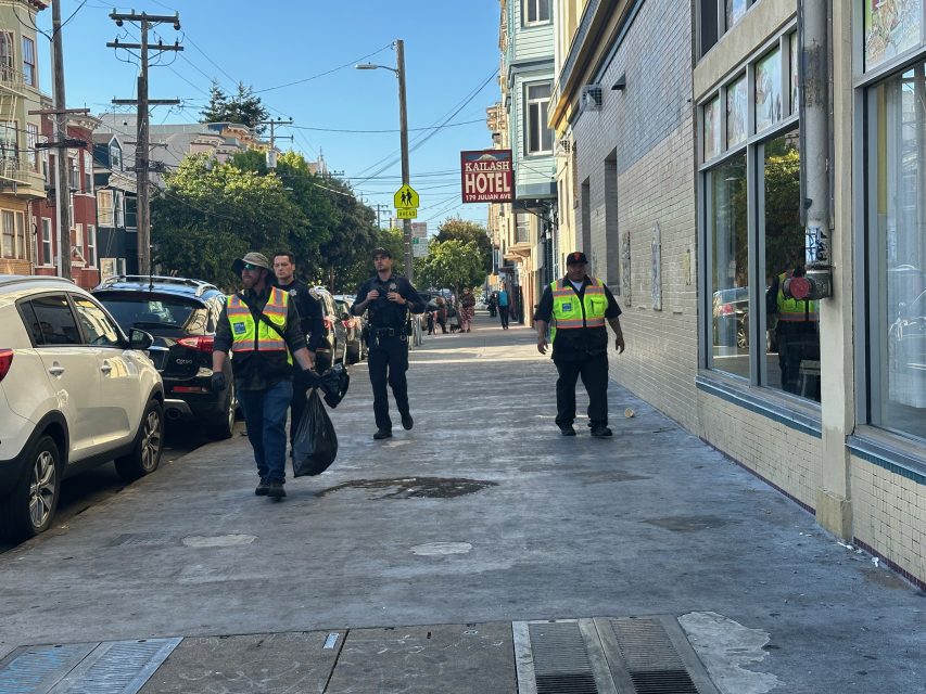 Three workers in reflective vests walk down a city sidewalk carrying trash bags; a police officer walks behind them.