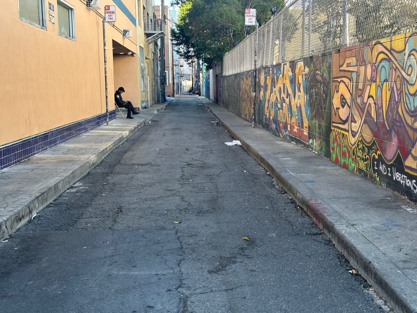 A narrow urban alleyway with graffiti-covered walls on the right, a person sitting on a bench to the left, and street signs visible above.