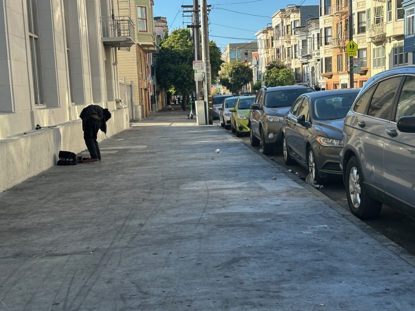 A person bends over near a wall on a wide city sidewalk lined with parked cars and apartment buildings on a clear day.