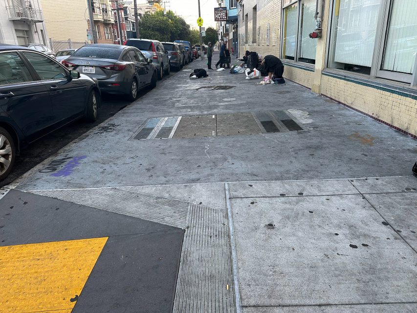 People are sitting and sorting belongings on a city sidewalk near parked cars, with buildings and a hotel visible in the background.