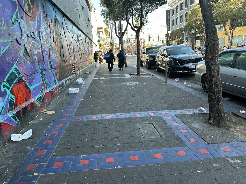 A city sidewalk with scattered litter, a painted mural on the left wall, and a few people walking in the distance next to parked cars.