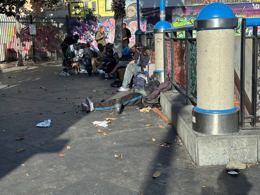 A group of people sit and lie on a city sidewalk surrounded by litter, with graffiti-covered walls and metal fencing in the background.