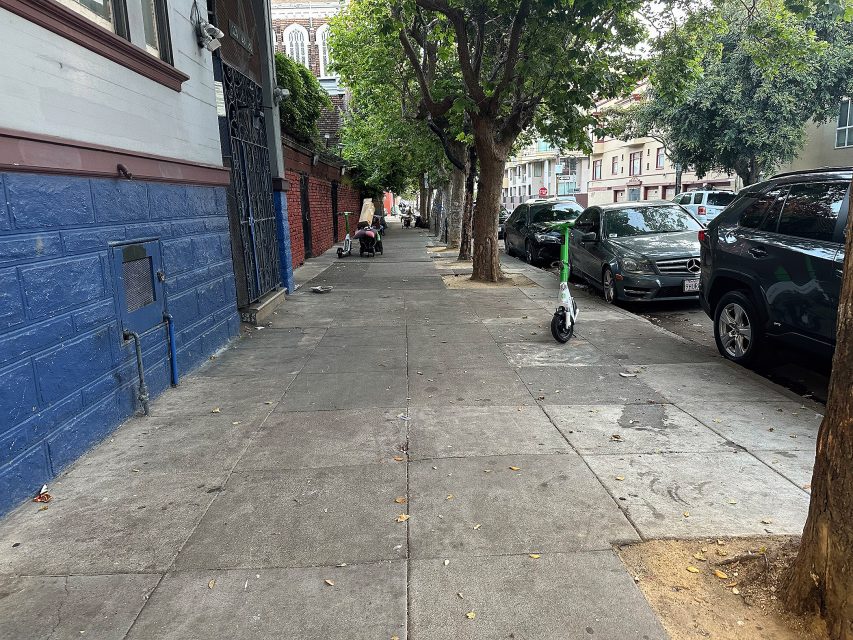 A sidewalk lined with trees and parked cars, with a green electric scooter and a person pushing a stroller in the distance.