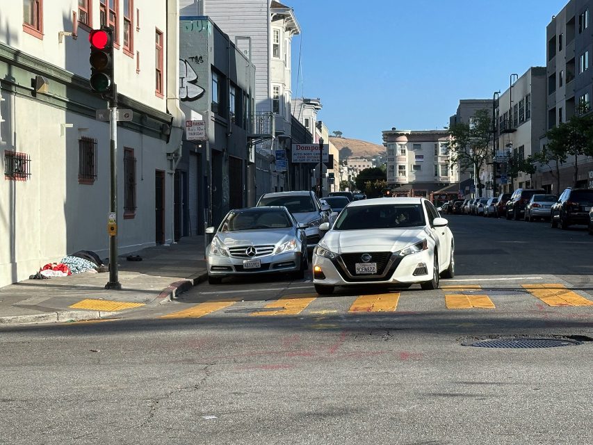 Two cars are parked across a crosswalk at an intersection in an urban area, blocking pedestrian access. The traffic light is red, and the street is mostly empty.
