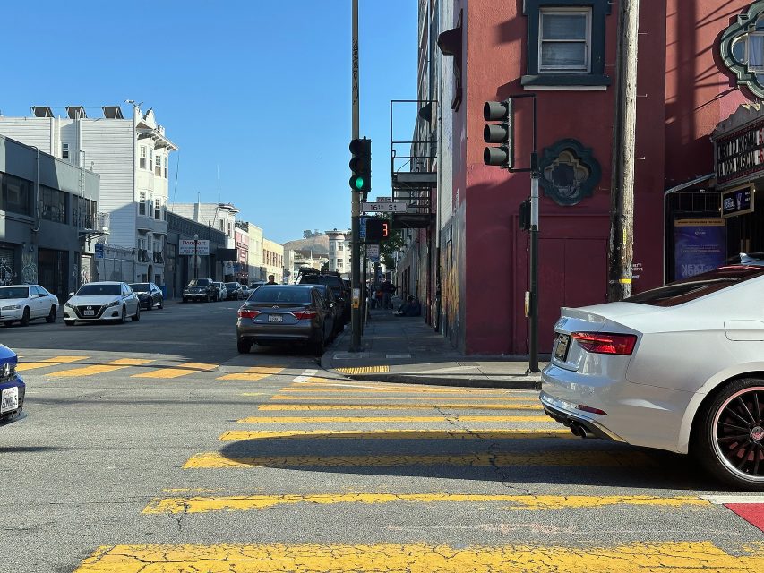 Intersection with yellow crosswalk lines, cars stopped at a red light, and buildings lining both sides of the street under a clear sky.