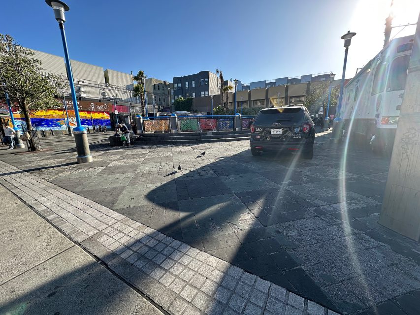 A city plaza with people sitting on benches, a police SUV parked nearby, colorful murals on a building, and a bus on the right, under a clear blue sky.