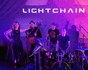 Four musicians stand and sit with instruments on a stage under purple lighting, with "LIGHTCHAIN" displayed on the wall behind them.