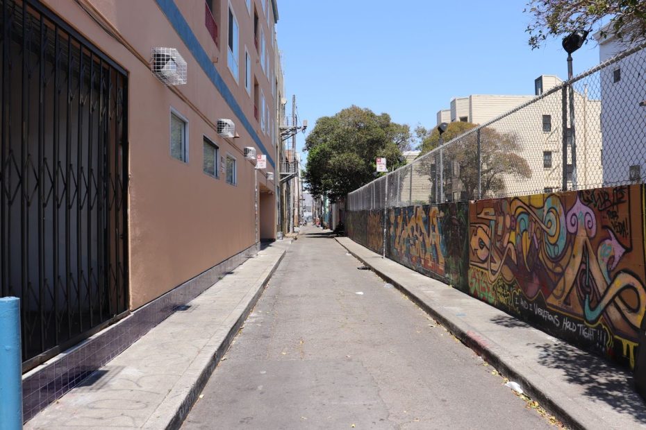 A narrow urban alleyway with graffiti on a fence to the right and apartment buildings on the left under a clear sky.
