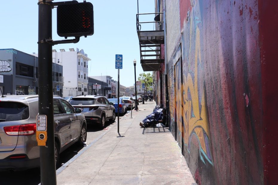 A city sidewalk with parked cars, a crosswalk signal, trash bags, and a building covered in graffiti art under a clear sky.