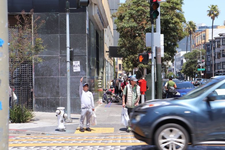 A child stands on a city sidewalk pointing upward while people and cars move through a busy urban intersection on a sunny day.