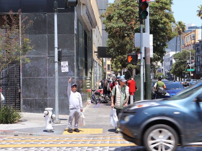 A child stands on a city sidewalk pointing upward while people and cars move through a busy urban intersection on a sunny day.