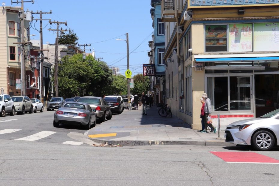 A city street corner with parked cars, pedestrians on the sidewalk, a hotel sign, and storefronts on a sunny day.