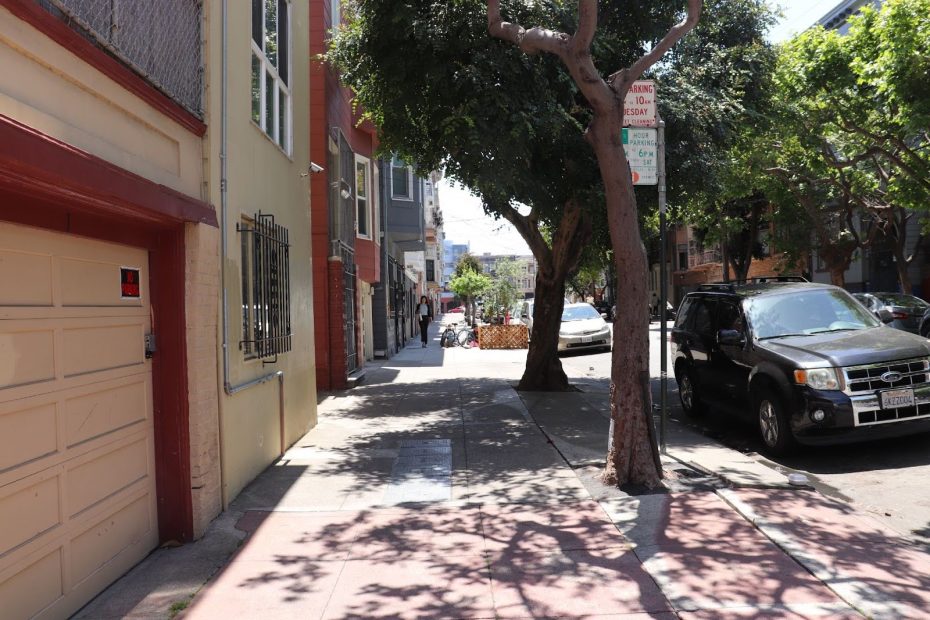 Sunny city sidewalk with trees, parked cars, and a beige garage on the left. A street sign and shadows are visible.