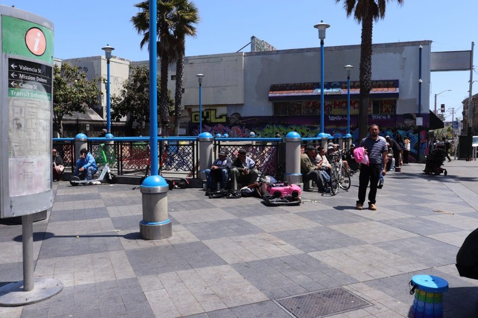 A city plaza with several people sitting near a railing, some with wheelchairs; a few pedestrians walk by; buildings and graffiti are in the background.