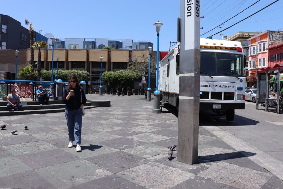 A woman walks through a city plaza near a white mobile command vehicle; other people are sitting on benches and pigeons are on the ground.