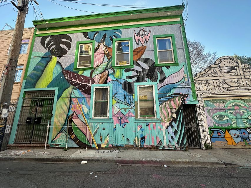 A two-story building with green-trimmed windows covered in colorful, abstract mural art, located on an urban street with street art on the adjacent wall.