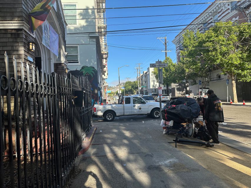 A white pickup truck is parked on a city street near a crosswalk as two people stand beside a shopping cart filled with belongings. Buildings and power lines are visible in the background.