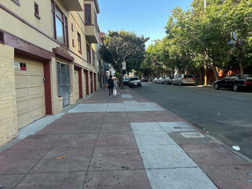 A person walks a dog on a mostly empty sidewalk beside parked cars and residential buildings on a tree-lined street.