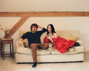 A man in casual clothes and a woman in a red ruffled dress sit on a beige couch in a simply decorated room with wooden beams.