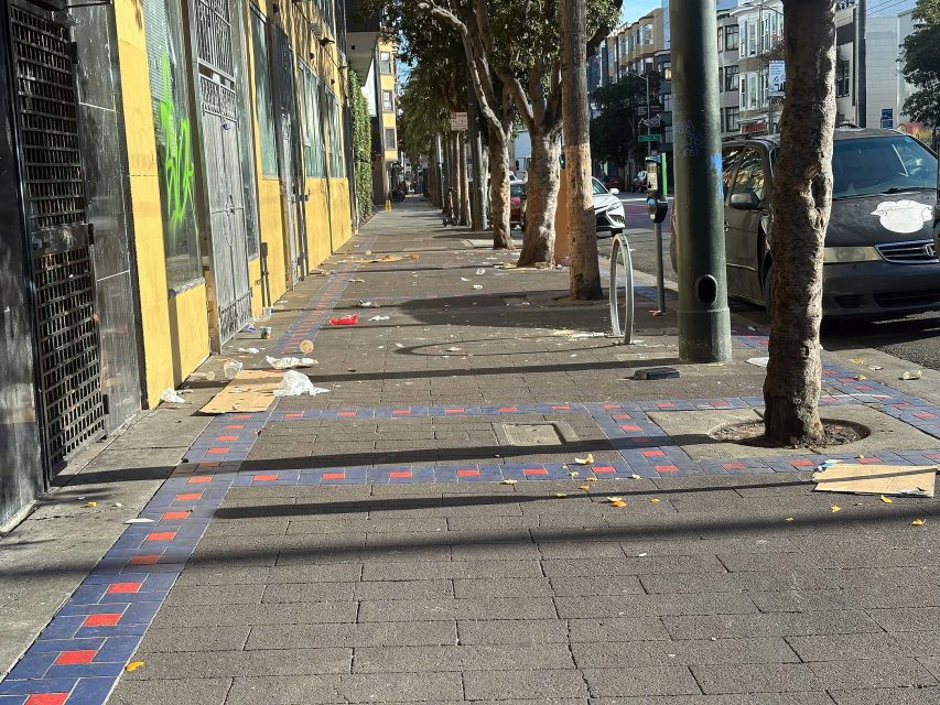 A city sidewalk lined with trees, scattered litter, parked cars, and buildings on one side on a sunny day.