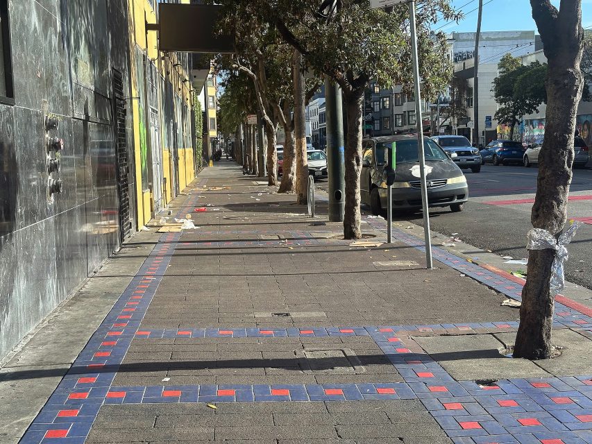 A city sidewalk with patterned tiles, litter scattered along the walkway, trees lining the side, and parked cars on the adjacent street during daylight.