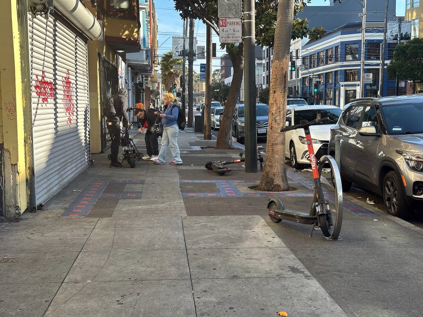 Two electric scooters lie on a city sidewalk near parked cars; three people with strollers stand and talk nearby under a "No Parking" sign.