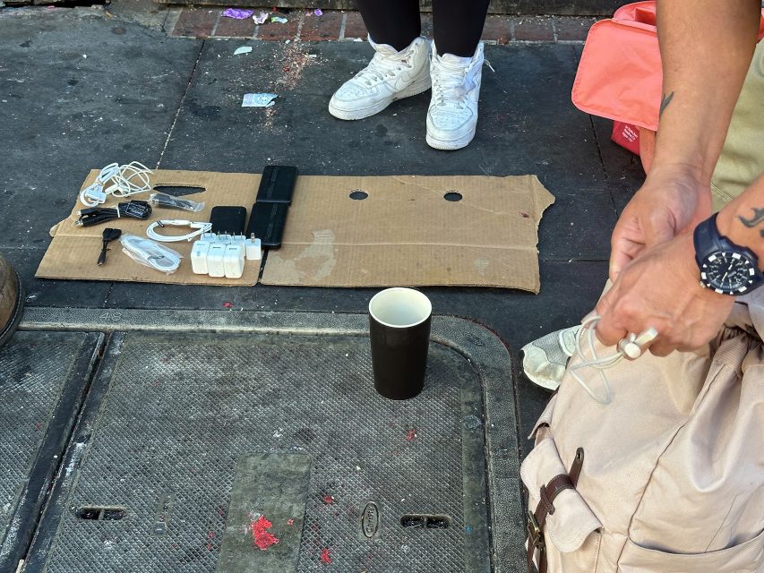 View of a cardboard mat on a sidewalk displaying various electronic chargers and cables, with a black paper cup and people standing nearby.
