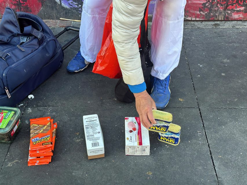 A person in white clothing arranges food items, including canned Spam, crackers, and snacks, on a sidewalk next to a duffel bag and a red plastic bag.