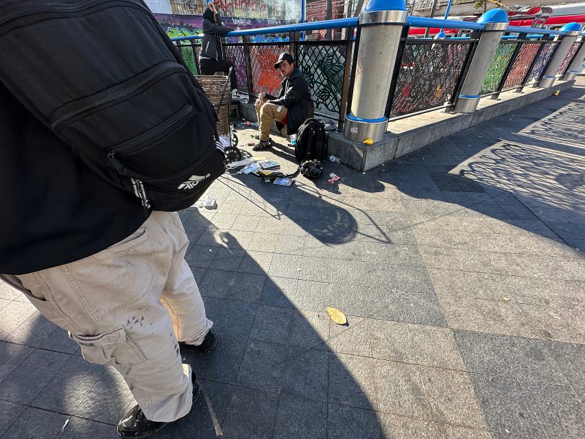 A man sits on the ground by a fence surrounded by belongings, while another person stands nearby on a sunlit city sidewalk.