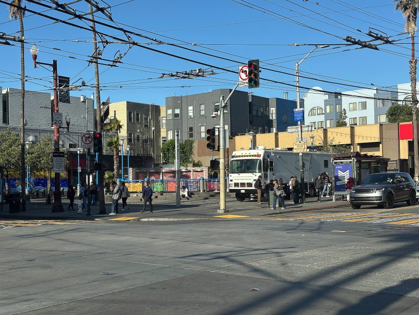 A city intersection with pedestrians crossing, a large white utility vehicle parked by the curb, and buildings in the background under clear blue skies.
