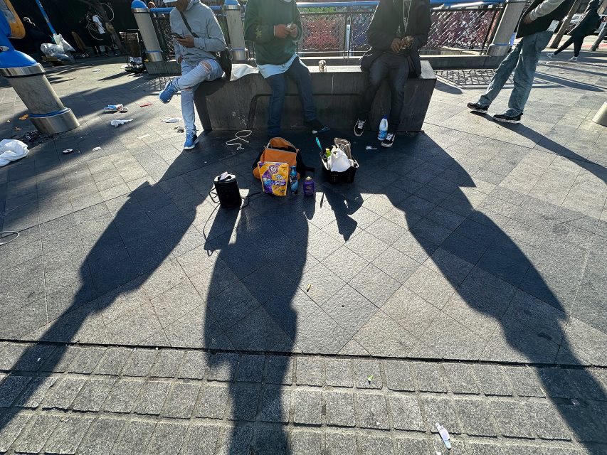 Three people sit on a concrete bench in sunlight, with shadows cast on the ground and assorted bags, drinks, and food items placed between them. Trash is scattered nearby.