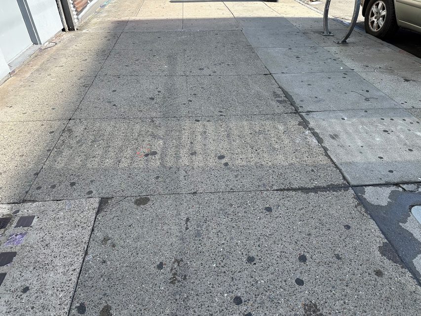 A worn and stained city sidewalk with faint outlines of where a crosswalk used to be; part of a parked car and bike rack are visible on the right.