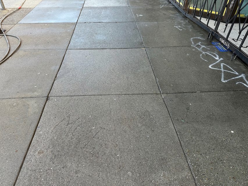 Concrete sidewalk with wet and dry areas, some faint graffiti, a hose on the left, and a black metal fence on the right.