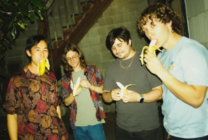 Four young men stand together near stairs, each holding and eating a peeled banana.
