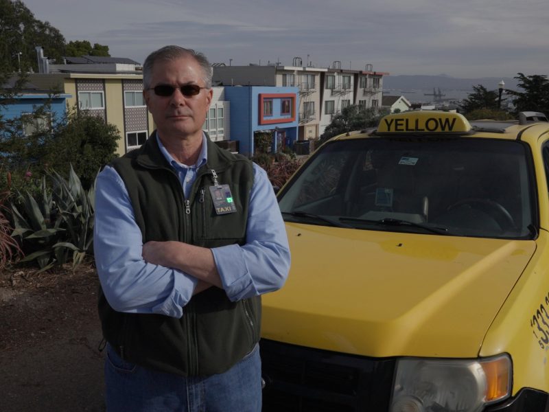 A man wearing sunglasses and a vest stands with arms crossed in front of a yellow taxi parked near residential buildings.